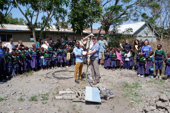 1 Spendenübergabe am Bohrloch für den Brunnen der Lekrimuni Primary School Eine Schulklasse steht um zwei Männer. Sie sehen alle sehr glücklich aus. Die Männer übergeben sich gegenseitig einen Spendencheck.