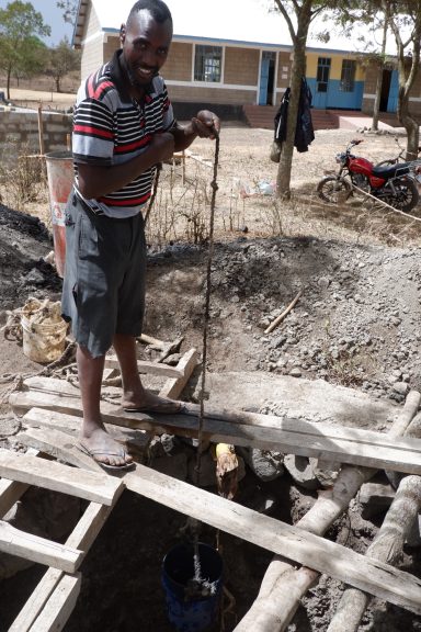 5 Arbeiten am Bohrloch mit... Ein Mann steht über einem Gerüst aus ein paar Latten mit einem Eimer an einem Seil und lässt diesen in das Loch ab.